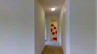 This single-floor residential space is thoughtfully arranged to maximize functionality and ease of movement. The interior hallway serves as a central corridor, connecting the various rooms of the home with a clean, straightforward layout. The hallway is finished with neutral, light-toned walls and carpeted flooring in a warm beige hue, contributing to a cozy and inviting ambiance throughout.

A flush-mount ceiling light fixture provides consistent overhead illumination along the hallway, ensuring the passage remains bright and welcoming. At the end of the corridor, a bathroom is visible, featuring a vibrant floral-patterned shower curtain in bold red and white tones that adds a lively pop of color to the otherwise understated palette. A small accent rug coordinates with the curtain near the bathroom entry.

To the side of the hallway, a louvered bi-fold closet door offers convenient built-in storage, a practical feature that enhances the utility of the space. The clean white trim and door frames throughout provide a crisp, finished look that ties the design together cohesively.

Overall, this space presents a functional and neatly maintained single-level layout with neutral finishes, adequate lighting, and practical storage solutions, making it a comfortable and efficient place to call home.