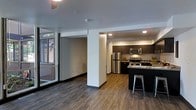 This contemporary single-floor space showcases a thoughtfully designed open-concept layout that seamlessly connects the living and kitchen areas. The kitchen is a standout feature, outfitted with dark espresso cabinetry, stainless steel appliances including a refrigerator, microwave, and range, and a breakfast bar with seating — ideal for casual dining or entertaining. The countertops provide ample workspace, while recessed lighting overhead keeps the area well-lit and functional. Rich, dark wood-style hardwood flooring runs throughout the space, adding warmth and a sense of continuity to the open floor plan. A structural column adds an architectural element that subtly defines the transition between the living and kitchen zones. Large floor-to-ceiling windows along one wall flood the living area with natural light and offer views of the surrounding greenery, creating a bright and airy atmosphere. The clean white walls and modern finishes throughout contribute to a minimalist yet inviting aesthetic. The overall ambiance is sleek, functional, and contemporary, making this a well-appointed space suited for modern living.