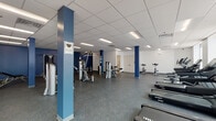 This fitness center offers a spacious, single-floor layout designed to accommodate a full range of exercise activities. The open floor plan is anchored by bold blue structural columns that add a distinctive visual element against the crisp white walls and drop-tile ceiling with recessed fluorescent lighting. Durable speckled rubber flooring runs throughout the main workout area, providing both safety and comfort underfoot.

The cardio section is well-appointed with a row of Spirit-brand treadmills and additional Spirit cardio machines, positioned near large windows that bring in ample natural light and create an airy atmosphere. Stationary spin bikes are also available for cycling workouts. The strength training area features a variety of cable machines, weight benches, a multi-function cable tower, and a vertical knee raise station, supported by medicine balls and resistance tools for functional training.

A water fountain and waste receptacle are conveniently located within the workout space. The facility also includes an accessible restroom equipped with grab bars and wood-look tile flooring, ensuring compliance with accessibility standards. The overall ambiance is clean, functional, and professional, making this fitness center a practical and well-maintained environment suited for a wide range of fitness needs.