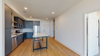 This single-floor unit showcases a thoughtfully designed open-concept layout that seamlessly connects the kitchen and living areas. The kitchen is a standout feature, outfitted with slate-blue cabinetry, stainless steel appliances including a French door refrigerator, electric range, and dishwasher, complemented by a subway tile backsplash and light wood floating shelves. A center island with a white countertop and blue metal frame provides additional prep space and casual seating. Light wood-tone flooring flows throughout the main living area, enhancing the bright, clean aesthetic created by white walls and recessed lighting overhead.

The living space is generously proportioned, offering an open canvas with ample room for flexible furniture arrangements. The bedroom features soft wall-to-wall carpeting, a ceiling fan, and a large window that fills the room with natural light while offering outdoor views. The bathroom is finished with large-format tile flooring, a walk-in shower, a vanity with a white countertop and gray cabinetry, and built-in open shelving for storage. The overall design is contemporary and cohesive, with neutral tones and modern finishes throughout.