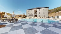 This expansive outdoor amenity space centers around a generously sized swimming pool with crystal-clear blue water, designed to evoke a resort-like atmosphere. The surrounding deck is finished with a striking two-tone checkered tile pattern, lending a polished and upscale character to the space. Multiple wicker chaise lounge chairs are arranged along the pool's perimeter, offering ample seating for relaxation and sunbathing beneath the open sky. Two wooden pergolas frame the deck area, providing shaded gathering spots and enhancing the overall aesthetic with a warm, natural contrast to the light-toned hardscape. A water feature adds a dynamic element to the pool's surface, catching sunlight and contributing to the lively ambiance. The pool area is enclosed by ornamental fencing, ensuring privacy and safety, while the surrounding residential buildings — finished in neutral gray siding with shuttered windows and balconies — create a cohesive community backdrop. Natural hillside scenery is visible beyond the fence line, adding a scenic, verdant boundary to the space. A dedicated clubhouse-style structure with large glass doors, covered porticos, and a distinctive roofline anchors one end of the pool, seamlessly connecting indoor and outdoor living. The overall environment is bright, open, and thoughtfully designed for leisure and community enjoyment.