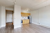 Located in Worcester, MA, this efficiently designed single-floor unit makes smart use of its compact footprint. The layout flows from a narrow hallway into an open combined living, dining, and kitchen area that forms the heart of the space. Light wood-tone laminate flooring runs consistently throughout, lending a clean, cohesive look. The living and dining area is bright, featuring two windows that frame views of the surrounding cityscape, complemented by a ceiling fan overhead and baseboard heating beneath the sills. The kitchen is neatly arranged along one wall, outfitted with warm honey-toned wood cabinetry — both upper and lower — paired with a white subway tile backsplash accented by bold red and black square tile details. Stainless steel appliances, including a refrigerator and a range, complete the functional kitchen setup. The bathroom continues the tile theme with floor-to-ceiling white subway tiles, again punctuated by matching red and black accent squares, and includes a full bathtub and shower combination alongside a vanity with sink. A closet and a utility room provide practical storage solutions. The unit's neutral gray-white walls, consistent flooring, and natural light from city-facing windows create a bright, functional living environment well-suited to urban living in downtown Worcester.