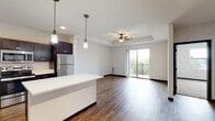 This well-designed single-floor residence offers a thoughtful layout that balances open communal spaces with comfortable private quarters. The heart of the home is a spacious open-concept kitchen and living area, unified by rich hardwood-style flooring throughout. The kitchen is a standout, featuring dark espresso cabinetry, crisp white quartz countertops, a large central island with pendant lighting, and a full suite of stainless steel appliances including a refrigerator, range, microwave, and dishwasher. A tile backsplash adds a clean, modern touch. The living area flows naturally from the kitchen and is highlighted by a tray ceiling with a ceiling fan, recessed lighting, and a sliding glass door that opens to a private balcony with outdoor views. A secondary living or flex space features track lighting and continues the warm hardwood-style flooring. The bedrooms are carpeted for comfort, with one featuring a tray ceiling, ceiling fan, and a large window that fills the room with natural light. The bathroom is finished with light tile flooring, dark vanity cabinetry, a soaking tub, and shaker-style interior doors, contributing to the clean, contemporary aesthetic found throughout the entire residence.