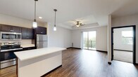 This well-designed single-floor residence offers a thoughtful layout that balances open communal spaces with comfortable private quarters. The heart of the home is a spacious open-concept kitchen and living area, unified by rich hardwood-style flooring throughout. The kitchen is a standout, featuring dark espresso cabinetry, crisp white quartz countertops, a large central island with pendant lighting, and a full suite of stainless steel appliances including a refrigerator, range, microwave, and dishwasher. A tile backsplash adds a clean, modern touch. The living area flows naturally from the kitchen and is highlighted by a tray ceiling with a ceiling fan, recessed lighting, and a sliding glass door that opens to a private balcony with outdoor views. A secondary living or flex space features track lighting and continues the warm hardwood-style flooring. The bedrooms are carpeted for comfort, with one featuring a tray ceiling, ceiling fan, and a large window that fills the room with natural light. The bathroom is finished with light tile flooring, dark vanity cabinetry, a soaking tub, and shaker-style interior doors, contributing to the clean, contemporary aesthetic found throughout the entire residence.