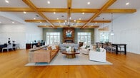 Located at 3795 Bismarck Ct in Wildwood, Florida, this single-story community clubhouse is designed with a modern farmhouse aesthetic that balances warmth and functionality. Wide-plank hardwood flooring flows continuously throughout the space, complementing the exposed natural wood beam ceilings that define the architectural character of the interior.

The main gathering area serves as the centerpiece of the clubhouse, anchored by a floor-to-ceiling stacked stone fireplace with a linear electric insert. Multiple seating arrangements, including leather sofas and swivel accent chairs, create inviting conversation areas beneath coffered, beam-detailed ceilings fitted with recessed lighting and contemporary chandeliers. French doors along the perimeter provide abundant natural light and direct access to outdoor amenities, including a visible pool area.

A dedicated reception and leasing office features pendant lighting, wainscoting, and a welcoming desk area with informational displays. A games and lounge area includes space for recreational activities such as foosball, alongside comfortable casual seating. A bar-height dining counter and additional lounge zones further extend the social areas of the space. Throughout, oversized windows and transomed French doors ensure the interior remains bright, airy, and visually connected to the outdoors.