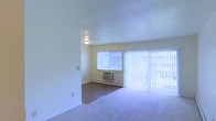 This single-floor space features an open living area characterized by clean, neutral-toned walls and soft carpet flooring throughout. The room is brightened by two sources of natural light: a standard window fitted with a wall-mounted air conditioning unit beneath it, and a large sliding glass door dressed with full-length vertical blinds, which together allow ample daylight to fill the interior. The sliding door provides potential access to an outdoor area while contributing to the room's airy feel. A flush-mount ceiling light fixture provides overhead illumination, and a baseboard heating unit runs along the lower wall beneath the window, indicating a dual-function climate control setup. Electrical outlets and a light switch are visibly positioned along the walls, reflecting practical, functional design. The overall layout is straightforward and open, offering a blank canvas well-suited for flexible furniture arrangements. The pale walls and light carpeting create a neutral, cohesive aesthetic throughout the space.
