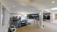 This fitness center offers a spacious, single-floor layout designed to accommodate a comprehensive workout experience. The facility is organized into distinct training zones, including a dedicated free weights area stocked with a full dumbbell rack on rubber matting, and an expansive cardio section lined with treadmills and upright cycling machines, each equipped with individual flat-screen monitors for entertainment during workouts. Additional strength training equipment is visible throughout the space, providing ample options for a full-body regimen. The interior features a neutral color palette with warm beige tones, recessed lighting throughout the ceiling, and decorative coffered ceiling details that lend the space a polished, upscale feel. Structural columns are integrated seamlessly into the layout, dividing the space into functional areas without disrupting the overall open feel. Large mirrors along the walls enhance the sense of depth and allow for proper form monitoring during exercise. Wall-mounted sconces add accent lighting, complementing the bright recessed fixtures overhead. A glass-panel entry door at the far end allows natural light to filter in, contributing to an inviting and energizing atmosphere suited for daily fitness activity.