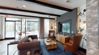 This thoughtfully designed single-floor space masterfully blends industrial and contemporary aesthetics across several distinct functional areas. The lounge area anchors the space with exposed dark wood ceiling beams, rich hardwood flooring, and a bold blue area rug beneath a tufted sofa and leather wingback chairs. A large wall-mounted flat-screen TV is framed by artistic brushstroke wallpaper murals, while floor-to-ceiling glass doors flood the room with natural light and provide direct access to an outdoor patio. The game room features a full-size pool table set beneath a soaring ceiling with an industrial steel staircase and mezzanine railing overhead. Built-in shelving with accent lighting lines the wall beneath the staircase, creating a cozy reading nook with lounge seating. A wet bar with bar stool seating and an additional mounted TV completes this entertainment hub. The bar kitchen area showcases a substantial marble-topped island with black tile detailing, industrial pendant lighting, open brass shelving against a white subway tile backsplash, and dark cabinetry. A dedicated dining nook features tufted leather banquette seating, marble-topped tables, brass pendant lighting, and the same signature brushstroke wall mural, lending the entire space a cohesive and sophisticated character.