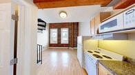 Located at 2018-2030 University Ave in Berkeley, CA, Unit 6 is a two-story loft-style residence that blends industrial character with functional living. The main floor hosts an open-plan kitchen, dining, and living area, where light hardwood flooring runs throughout and a striking exposed brick wall serves as a defining architectural feature. The kitchen is equipped with wood cabinetry, granite-style countertops, a gas range, built-in microwave, and a full-size refrigerator. Wooden beam accents along the ceiling add warmth and texture to the space, while windows facing the exterior bring in natural light. A full bathroom on the main level features a combined tub and shower with sliding glass doors and tile flooring. A dramatic spiral staircase with a black metal railing connects the main floor to the upper loft level. The loft is a bright, airy retreat highlighted by soaring vaulted ceilings with exposed wood beams and a large skylight that floods the space with natural light. Exposed brick continues here, reinforcing the unit's industrial aesthetic. The loft includes a closet with mirrored sliding doors and open railing overlooking the lower level, creating a sense of openness and visual connection between floors.