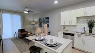 This single-story residence showcases an open-concept layout that integrates the kitchen and living areas into one cohesive, functional space. The kitchen is outfitted with crisp white cabinetry featuring sleek hardware, a classic white subway tile backsplash, and stainless steel appliances, including a dishwasher. A white kitchen island serves as a central gathering point, offering casual seating with bar-style stools and a clean surface ideal for dining or meal prep. Recessed lighting illuminates the kitchen area, while a ceiling fan with an integrated light fixture provides both style and comfort to the adjacent living space. The living area is finished with neutral gray walls that create a calm, versatile backdrop, complemented by a patterned area rug that anchors the seating arrangement. A floor lamp adds warm ambient lighting, and a framed piece of wall art provides a focal point. Natural light filters into the space through a draped window, enhancing the overall bright and airy atmosphere. The cohesive neutral palette throughout gives the space a modern, welcoming character.