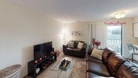 This single-floor residence offers a practical and comfortable layout designed to accommodate multiple occupants. The living area serves as the social hub of the unit, featuring plush carpeting, a leather sofa and loveseat arrangement, a glass-top coffee table, and a wall-mounted television setup — all bathed in warm, ambient lighting. A tiled entryway connects seamlessly to the main living space, creating a natural transition point upon entry.

The kitchen is efficiently designed with light-toned cabinetry, tile flooring, a full-size refrigerator, and standard appliances including a range and dishwasher, providing all essential cooking amenities in a compact footprint.

The unit includes multiple bedrooms, each carpeted and finished with neutral walls and textured ceilings. One bedroom features a dedicated study area with a desk, a corkboard for organization, a wall-mounted mirror, and ample floor space. A second bedroom includes a window with blinds and curtains, a closet, and room for both sleeping and study furniture.

Two full bathrooms serve the unit, each equipped with a tub-shower combo fitted with sliding glass doors, a toilet, vanity, and towel hardware — offering functional and private bathing spaces for all residents.