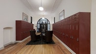 Located at 345 East Deshler Avenue in Columbus, Ohio, this thoughtfully preserved historic building seamlessly blends original architectural character with modern functionality. The space occupies a single floor and makes a strong first impression with its corridor featuring rows of deep burgundy metal lockers lining both walls — a nod to the building's storied past. Warm-toned hardwood flooring runs throughout, complementing the rich color palette of the lockers and lending a grounded, classic feel underfoot. The hallway opens to a casual seating arrangement of leather club chairs gathered around a low table on a plaid area rug, creating an inviting gathering spot. Above, ornate crown molding and high ceilings with globe pendant lights enhance the sense of grandeur while maintaining an approachable warmth. At the far end, a striking arched window with black steel framing floods the space with natural light, serving as a dramatic focal point. Framed artwork adorns the walls at intervals, adding a curated, thoughtful touch to the overall aesthetic. The combination of historic detail, repurposed industrial elements, and comfortable furnishings gives this Columbus property a distinctive character that honors its architectural heritage.