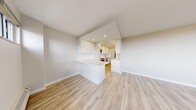 This well-appointed single-floor unit offers a clean, modern aesthetic defined by light hardwood flooring, neutral white and warm beige walls, and an abundance of natural light throughout. The open-concept living and kitchen area forms the heart of the space, where a peninsula counter creates a natural divide between the two zones. The kitchen is outfitted with stainless steel appliances — including a range, over-the-range microwave, and dishwasher — complemented by crisp white cabinetry, white quartz-style countertops, and a subway tile backsplash. The living area is generously sized and flows naturally toward a wall of large floor-to-ceiling windows that frame expansive outdoor views of treetops and the surrounding neighbourhood, filling the room with daylight. A hallway connects the main living area to two well-proportioned bedrooms, each featuring double closets with panelled white doors. One bedroom includes convenient access to a dedicated in-unit laundry closet housing a stacked washer and dryer. The full bathroom is finished in an all-white palette, complete with a vanity, mirror, toilet, and a soaker tub with tiled surround. Throughout the unit, clean lines and consistent finishes create a cohesive, move-in-ready environment.