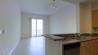 This single-floor space offers an airy, open-plan layout that seamlessly connects the kitchen and living area, creating a sense of generous space and flow. The kitchen is equipped with rich granite countertops that wrap around a breakfast bar peninsula, a stainless steel sink, and a built-in dishwasher — providing both style and practicality for everyday living. Light-toned large-format tile flooring extends throughout the main living area, reflecting natural light and enhancing the clean, contemporary aesthetic. A track lighting fixture overhead adds a modern accent while ensuring the space is well-lit. The living area is bathed in natural light, thanks to a full-height sliding glass door that opens onto a private balcony. The balcony offers expansive views of the surrounding cityscape, making it a standout feature of the space. The interior walls are finished in crisp white, complementing the bright and open atmosphere. Vertical blinds along the balcony door allow for flexible light control and privacy. The overall ambiance is clean, modern, and bright, with an emphasis on open space and natural light throughout.