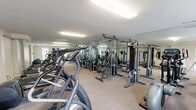 Located at 4573 Whisper Lake Dr in Black Jack, Missouri, this fitness center offers a compact yet thoroughly equipped workout facility on a single floor. The space is designed to maximize functionality, with floor-to-ceiling mirrors lining the walls to create an expansive, open feel and allow users to monitor their form throughout their workouts.

The cardio section features multiple treadmills and elliptical trainers, providing ample options for aerobic exercise. The strength training area is equally well-appointed, housing a variety of cable machines, lat pulldown stations, chest press rigs, and adjustable benches — offering a comprehensive range of resistance-based workout options. Additional stationary bikes round out the cardio equipment selection.

Neutral-toned carpet covers the floor throughout, providing cushioning underfoot and a clean, polished appearance. Recessed fluorescent lighting ensures the space is brightly and evenly illuminated. The white walls and mirrored surfaces work together to amplify the sense of space, making the gym feel larger than its footprint suggests. Ventilation is provided through ceiling vents, maintaining a comfortable environment during exercise. Overall, this fitness center is a well-organized and fully functional facility suited for a broad range of fitness routines.