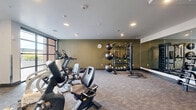 Located at 4277 S Main St in Murray, Utah, this fitness center offers a well-equipped exercise space designed to support a comprehensive workout routine. The room features a neutral color palette with olive-toned accent walls, light gray carpeted flooring, and recessed lighting throughout, creating a clean and focused environment for training.

Cardio equipment includes treadmills, elliptical machines, and stationary bikes positioned near large floor-to-ceiling windows that allow abundant natural light and provide views of the surrounding outdoor area. A wall-mounted flat-screen television is conveniently positioned for entertainment during workouts.

The strength training area is well-stocked with a full rack of dumbbells, barbells, and a multi-functional rig equipped with resistance bands, medicine balls, and battle ropes for functional fitness training. A large wall-to-wall mirror spans one side of the room, visually expanding the space and allowing users to monitor their form during exercise.

The facility also includes a wall clock, a waste receptacle, and direct access to an outdoor area through a glass-paneled door. The overall layout is thoughtfully organized to provide ample open floor space for stretching or bodyweight exercises, making this fitness center a versatile and practical amenity.