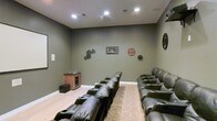 This single-floor home theater is thoughtfully designed to replicate the feel of a private cinema. The room is finished with deep olive-gray walls that absorb light and minimize glare, creating an ideal viewing environment. Recessed ceiling lights provide controlled, dimmable illumination, while the clean white crown molding and baseboards offer a subtle contrast to the darker palette.

Seating is arranged in two tiered rows of plush, dark leather power recliners, each equipped with built-in cup holders, ensuring comfort during extended viewing sessions. The elevated rear row provides unobstructed sightlines to the screen, mimicking a true theatrical layout. A patterned area rug anchors the floor between the rows, adding warmth and texture to the light hardwood flooring.

Cinema-themed wall décor, including vintage film reel pieces and gear motifs, reinforces the room's dedicated purpose and adds character. A wooden media cabinet with a glass-front wine or beverage cooler is neatly positioned along one wall, offering convenient in-room refreshment storage. A subwoofer placed nearby suggests a capable surround sound system. Overall, the space balances functional design with an inviting, entertainment-focused ambiance.