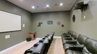 This single-floor home theater is thoughtfully designed to replicate the feel of a private cinema. The room is finished with deep olive-gray walls that absorb light and minimize glare, creating an ideal viewing environment. Recessed ceiling lights provide controlled, dimmable illumination, while the clean white crown molding and baseboards offer a subtle contrast to the darker palette.

Seating is arranged in two tiered rows of plush, dark leather power recliners, each equipped with built-in cup holders, ensuring comfort during extended viewing sessions. The elevated rear row provides unobstructed sightlines to the screen, mimicking a true theatrical layout. A patterned area rug anchors the floor between the rows, adding warmth and texture to the light hardwood flooring.

Cinema-themed wall décor, including vintage film reel pieces and gear motifs, reinforces the room's dedicated purpose and adds character. A wooden media cabinet with a glass-front wine or beverage cooler is neatly positioned along one wall, offering convenient in-room refreshment storage. A subwoofer placed nearby suggests a capable surround sound system. Overall, the space balances functional design with an inviting, entertainment-focused ambiance.