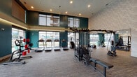 Located at 14175 Dallas Pkwy in Farmers Branch, TX, this well-appointed fitness center offers a comprehensive, single-floor wellness facility designed with both function and aesthetics in mind. The main workout area is expansive and filled with natural light streaming through floor-to-ceiling windows, which frame views of the surrounding landscape. Bold teal accent walls, textured stone-patterned surfaces, and elevated ceilings create a dynamic, modern atmosphere throughout the space.

The primary gym floor is equipped with a wide array of cardio machines, including treadmills, ellipticals, a stair climber, and rowing machines, all positioned to take advantage of the generous window views. Strength training is well-supported with a multi-station cable machine, selectorized weight equipment, free weights, and plyo boxes for high-intensity workouts.

A dedicated studio room features rich hardwood flooring, full-length mirrors, large windows, and a ceiling fan, making it ideal for yoga, group fitness classes, or personal training sessions.

A lounge area adjacent to the gym includes a striking wood-panel accent wall, a wall-mounted flat-screen TV, built-in cabinetry, and a mini-refrigerator, providing a welcoming space to relax before or after a workout. Recessed lighting and carefully considered finishes tie the entire facility together into a cohesive, premium fitness experience.