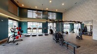 Located at 14175 Dallas Pkwy in Farmers Branch, TX, this well-appointed fitness center offers a comprehensive, single-floor wellness facility designed with both function and aesthetics in mind. The main workout area is expansive and filled with natural light streaming through floor-to-ceiling windows, which frame views of the surrounding landscape. Bold teal accent walls, textured stone-patterned surfaces, and elevated ceilings create a dynamic, modern atmosphere throughout the space.

The primary gym floor is equipped with a wide array of cardio machines, including treadmills, ellipticals, a stair climber, and rowing machines, all positioned to take advantage of the generous window views. Strength training is well-supported with a multi-station cable machine, selectorized weight equipment, free weights, and plyo boxes for high-intensity workouts.

A dedicated studio room features rich hardwood flooring, full-length mirrors, large windows, and a ceiling fan, making it ideal for yoga, group fitness classes, or personal training sessions.

A lounge area adjacent to the gym includes a striking wood-panel accent wall, a wall-mounted flat-screen TV, built-in cabinetry, and a mini-refrigerator, providing a welcoming space to relax before or after a workout. Recessed lighting and carefully considered finishes tie the entire facility together into a cohesive, premium fitness experience.