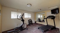 This single-floor fitness room is designed as a functional exercise space, offering a well-organized layout suitable for a variety of workout routines. The room is finished with durable, patterned carpet tiles in muted tones of burgundy and grey, providing a practical surface underfoot. Walls are painted in a soft yellow hue, complemented by white trim and a textured ceiling fitted with flush-mount light fixtures that provide consistent overhead illumination.

Natural light enters through multiple windows fitted with horizontal blinds, lending the space a bright, open feel. A large wall mirror is mounted along one side of the room, visually expanding the space and serving as a functional training aid. The room accommodates a treadmill, an elliptical trainer, and a multi-tier dumbbell rack stocked with a range of free weights, offering options for both cardio and strength training.

Access is provided through a set of white double doors with glass panels leading to the exterior, as well as an additional interior door connecting to an adjacent hallway. Signage including an exit sign and posted notices are present near the entry points. The overall layout is practical and uncluttered, making efficient use of the available space for fitness purposes.