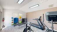 Located at 9400 Stagecoach Rd in Little Rock, Arkansas, this fitness center is a dedicated single-room exercise facility designed to accommodate a well-rounded workout experience. The space features vaulted ceilings that lend an open, airy feel despite the room's compact footprint, and is illuminated by large flush-mounted LED panel lights along with natural light streaming through multiple windows fitted with horizontal blinds. The patterned carpet flooring adds a practical and clean aesthetic throughout.

The room is outfitted with a comprehensive selection of exercise equipment, including a treadmill, elliptical trainer, stationary spin bike, and a weight bench. A full dumbbell rack stocked with a range of free weights lines one wall, complemented by a functional training station featuring medicine balls, resistance bands, and stability balls on a dedicated rack. A security camera is mounted near the ceiling, ensuring a monitored environment.

Large windows along one wall overlook the outdoor pool area, providing pleasant views and additional natural light. A glass-panel door offers direct access to the pool deck, seamlessly connecting the fitness center to the broader outdoor amenity space. The textured ceiling and neutral warm-toned walls complete the clean, functional atmosphere of this well-equipped community fitness room.