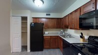 This single-floor residence offers a thoughtfully designed layout that balances functionality and comfort throughout its key living spaces. The kitchen is outfitted with rich medium-toned wood cabinetry providing generous storage, light-toned laminate countertops, and a full suite of black appliances including a refrigerator, range, microwave, and dishwasher. Wood-look tile flooring and a textured ceiling complete the practical yet welcoming feel of the space.

The living area is carpeted and bathed in natural light from multiple windows dressed with horizontal blinds, offering views of surrounding greenery. The adjoining dining area features a chandelier overhead and its own window, creating a pleasant setting for meals. Both spaces share warm neutral wall tones that contribute to a cohesive, relaxed ambiance.

The bedroom is generously sized with plush carpeting, multiple windows allowing ample natural light, and notable storage including both a walk-in closet and an additional standard closet. The bathroom is clean and bright, featuring wood-look tile flooring, a tub-shower combination, and white fixtures throughout. Overall, the unit presents a comfortable and practical living environment with consistent finishes across all areas.