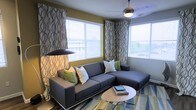 This single-floor space offers a thoughtfully designed interior that balances modern aesthetics with everyday comfort. The living area is a standout feature, anchored by a sleek, gray sectional sofa accented with white and green throw pillows that add pops of color against the neutral tones. A round, minimalist coffee table sits atop a patterned area rug in shades of blue, green, and gold, lending warmth and visual interest to the floor.

Large windows wrapped in patterned draping curtains flood the room with abundant natural light, creating an airy, open atmosphere while also offering views of the surrounding environment. The walls are painted in a warm olive-yellow tone, complementing the contemporary décor and adding depth to the space.

Overhead, a ceiling fan with a built-in light fixture provides both practical air circulation and subtle ambient lighting, while a modern arc floor lamp offers an additional layer of task lighting. The combination of layered lighting sources enhances the room's versatility for different times of day.

Overall, the space presents a cohesive, contemporary design with a carefully curated color palette, functional furnishings, and an inviting ambiance well-suited for relaxing or entertaining.
