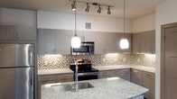 This single-story residence at Monarch in Austin's 78613 zip code offers a thoughtfully designed layout with contemporary finishes throughout. Dark wood-style plank flooring flows through the main living areas, complementing the neutral color palette and creating a cohesive, modern aesthetic.

The kitchen serves as a focal point of the home, featuring granite countertops, a central island with an undermount sink, flat-front cabinetry in a warm gray tone, stainless steel appliances including a range and built-in microwave, and a mosaic tile backsplash. Pendant lighting and track fixtures illuminate the space effectively.

The spacious living area opens directly onto a private balcony through sliding glass doors, allowing natural light to fill the room. The bedroom is carpeted for comfort and includes a generously sized walk-in closet outfitted with wire shelving systems for organized storage.

The bathroom features a soaking tub and shower combination with decorative tile accents, a granite-topped vanity, and wood-toned cabinetry. A dedicated laundry closet with full-size washer and dryer connections adds notable convenience to this well-rounded residence.