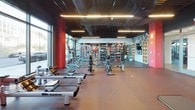 This fitness center offers a spacious, single-floor layout designed to accommodate a comprehensive range of workout activities. The interior is defined by a striking dark ceiling with exposed ductwork and sleek linear LED lighting, paired with bold red accent columns and walls that lend the space a dynamic, high-energy atmosphere.

The cardio zone is lined with treadmills, ellipticals, rowing machines, and stationary bikes, all positioned along floor-to-ceiling windows that provide abundant natural light and outdoor views, creating an open and motivating environment. A dedicated free weights area features adjustable benches, squat racks, and an extensive dumbbell rack displayed in front of full-length mirrors, allowing users to monitor their form effectively.

Strength training equipment and cable machines are distributed throughout the main floor, complemented by a heavy punching bag for functional training. A separate glass-enclosed room provides space for stretching or small group workouts. One wall features a bold graphic black-and-white art installation alongside built-in shelving and a mounted TV. Durable rubber and speckled flooring throughout supports both heavy lifting and cardio activity comfortably.