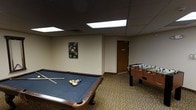 This single-floor recreational space is designed as a dedicated game and leisure room, offering a welcoming environment for entertainment and social gatherings. The room is anchored by a full-size pool table with a dark felt surface, complete with a racked set of billiard balls and cues, making it ready for play. Complementing it is a solid wood foosball table, adding variety to the recreational offerings. The flooring features a patterned carpet with an earthy, ornate design that lends a classic warmth to the space. Overhead, a drop ceiling with recessed fluorescent panel lighting provides even, practical illumination throughout the room. The neutral-toned walls are kept simple, accented by a single piece of decorative wall art, maintaining an uncluttered and functional atmosphere. A wood-paneled door leads to an adjacent area, suggesting this game room is part of a larger facility. Overall, the space strikes a balance between functionality and comfort, making it well-suited as a communal amenity for residents or guests looking to unwind and engage in friendly competition.