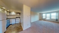 Located at 1600 Barnsdale Rd in La Grange Park, Illinois, this single-floor unit offers a straightforward, functional layout designed for comfortable everyday living. The open living area is generously proportioned, featuring neutral-toned carpeting that extends throughout the main living spaces and bedroom. A window unit provides cooling, while baseboard heating runs along the living room's exterior wall, ensuring year-round climate control. Natural light filters in through windows, lending a bright, airy feel to the open space.

The kitchen is set apart from the living area by a partial dividing wall, creating a semi-separated cooking space. It is equipped with white appliances including a refrigerator, electric range, and oven, complemented by gray cabinetry with glass-front upper cabinets and recessed lighting overhead. The flooring transitions to a durable stone-look tile in the kitchen area.

The bedroom offers a comfortable, carpeted space with a standard closet featuring a sliding door and built-in rod for storage. The full bathroom includes a tiled tub-shower combination, a wood-toned vanity with a mirror cabinet, tile flooring, and a baseboard heater. The overall design is clean and neutral, providing a practical, move-in-ready living environment in a residential La Grange Park setting.