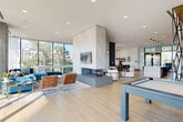 This contemporary amenity space showcases a thoughtfully designed open floor plan finished with wide-plank light hardwood flooring throughout. The expansive main lounge is anchored by a dramatic floor-to-ceiling double-sided fireplace clad in smooth concrete, surrounded by generous seating arrangements bathed in abundant natural light from large floor-to-ceiling glass walls that frame lush treetop views. A pool table adds a recreational focal point to the living area, while sleek steel-framed glass partitions elegantly define zones without sacrificing the sense of openness.

The gourmet kitchen features a substantial marble-waterfall island with bar seating, dark cabinetry extending to the ceiling, built-in appliances, and globe pendant lighting. A dedicated lounge nook offers a striking exposed concrete accent wall, wall-mounted display screen, and a circular statement chandelier, creating an intimate yet stylish retreat.

A formal meeting room is equipped with a large dark conference table, plush velvet seating, and pendant lighting, suitable for professional gatherings. Seamless access leads to a spacious covered outdoor terrace with exposed brick columns and a steel ceiling, furnished with lounge seating and dining sets. The resort-style pool deck features a shimmering pool, in-water lounge chairs, and open-air areas framed by the building's glass facade.