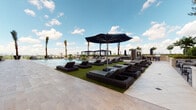 This rooftop amenity level presents a well-appointed outdoor retreat designed for relaxation and leisure. The deck is finished with warm-toned natural stone pavers that extend throughout the space, creating a cohesive and polished aesthetic. At the center of the amenity area is a resort-style pool surrounded by an expansive sun deck, where rows of dark wicker chaise lounges are arranged on lush synthetic turf panels beneath large navy blue market umbrellas. Multiple covered cabana structures line the perimeter, offering shaded alternatives for outdoor seating and gathering.

A dedicated lounge zone, situated adjacent to the building's glass curtain wall facade, features a curated arrangement of grey wicker armchairs and low-profile coffee tables framed by raised planters filled with manicured shrubs and ornamental grasses. These planted borders add a natural, verdant element to the otherwise contemporary hardscape. Floor-to-ceiling glazing reflects the open sky and surrounding cityscape, reinforcing the elevated, urban character of the space.

Access to the deck is provided through sleek aluminum-framed glass doors. The surrounding skyline is visible beyond the deck's perimeter, with palm trees accenting the outdoor landscape. The overall design balances modern architectural detailing with comfortable, resort-inspired furnishings.