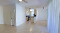 This single-floor space offers a clean, contemporary layout designed for comfortable everyday living. The open-plan design connects the living area and kitchen seamlessly, creating a sense of spaciousness and ease of movement throughout.

The living area features light hardwood flooring that extends across the entire unit, reflecting natural light and contributing to the bright, airy atmosphere. A ceiling fan overhead provides both function and a subtle design element. Track lighting adds additional illumination, ensuring the space is well-lit throughout the day and evening.

The kitchen is thoughtfully appointed with light wood cabinetry, dark countertops, and stainless steel appliances, including a range and microwave. A peninsula counter separates the kitchen from the living area, offering a practical transition between the two spaces while maintaining an open feel. A window in the kitchen allows natural light to filter in, enhancing the overall brightness of the space.

The white walls throughout the unit provide a neutral, versatile backdrop suitable for a variety of interior styles. Clean lines, consistent flooring, and an uncluttered layout make this a functional and welcoming space with a modern sensibility.