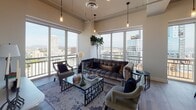 Located in the heart of Downtown Los Angeles, this contemporary residence at 225 S Olive St offers a sophisticated urban living experience defined by clean lines, premium finishes, and stunning panoramic city views. The open-concept layout flows seamlessly from room to room, beginning with a welcoming entry hall adorned with curated artwork and tile flooring. The chef's kitchen is outfitted with dual-tone cabinetry in grey and white, quartz countertops, stainless steel appliances including a built-in double oven and full-size refrigerator, a textured tile backsplash, and a central island with bar seating. The living and dining areas are bathed in natural light through expansive floor-to-ceiling windows and sliding glass doors that open onto private balconies, framing sweeping views of the Los Angeles skyline. The primary bedroom features plush carpeting, a ceiling fan, and direct balcony access, while the bathrooms are finished with large-format tile, glass-enclosed soaking tubs, separate shower enclosures, and illuminated vanity mirrors. A dedicated office or sitting room with wood-look flooring and gallery wall adds functional versatility to the space.