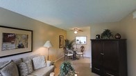 This single-floor residence offers a comfortable and functional layout with a seamlessly connected living and dining area. The living space is finished with light neutral walls that create a warm, inviting atmosphere, complemented by plush seating and accent lighting from a classic floor lamp. A textured popcorn ceiling adds a traditional character to the room, while tile flooring runs throughout, offering durability and ease of maintenance.

The dining area is situated adjacent to the living room, anchored by a round glass-top table surrounded by elegant wooden chairs. A ceiling fan with an integrated light fixture overhead provides both comfort and illumination. A window in the dining space allows natural light to filter in, brightening the area and creating an airy feel.

A large dark wood armoire serves as a striking focal point near the entryway, offering ample storage and a rich contrast against the lighter wall tones. Decorative greenery and framed wall art are thoughtfully placed throughout, adding personality and life to the space.

Overall, this residence presents a cohesive, well-appointed interior that balances practicality with a homely aesthetic, making it a comfortable and functional living environment.