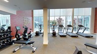 This well-appointed fitness center offers a thoughtfully designed workout environment suited for a wide range of exercise routines. The space is anchored by an impressive wall of floor-to-ceiling windows that flood the interior with natural light and provide inspiring views of an outdoor pool and surrounding landscaping, creating an energizing atmosphere for workouts.

The cardio zone is generously equipped with multiple treadmills, elliptical machines, and a stationary recumbent bike, all arranged to take full advantage of the outdoor views. An adjustable weight bench adds versatility for strength training. The neutral carpeted flooring provides comfort underfoot throughout the space.

A standout design feature is a bold accent wall in a deep rose hue, adorned with a topographic pattern and the motivational phrase "Great Things Never Came from Comfort Zones" in gold lettering, adding personality and energy to the environment. Structural columns are cleanly finished, complementing the modern aesthetic of the space.

The single-floor layout ensures easy accessibility, and the open, airy design allows for ample circulation between equipment. Overall, this fitness center combines functional amenities with an inviting, contemporary design to deliver an engaging and motivating workout experience.