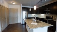 Located at 35 Folly Road Blvd in Charleston, South Carolina, this single-floor residence combines modern design with functional living. The kitchen serves as a standout centerpiece, showcasing rich dark cabinetry that extends to the upper walls, offering generous storage throughout. A large granite-topped island with an undermount sink anchors the space, providing both a practical workspace and a natural gathering point. Stainless steel appliances, including a full-size French door refrigerator and a dishwasher, lend a sleek, contemporary feel. A tiled mosaic backsplash adds texture and visual interest, complementing the overall color palette. Pendant lighting hangs above the island, while a track lighting system along the ceiling ensures the space is well-lit throughout. Dark hardwood flooring flows through the kitchen and into the adjoining areas, creating a cohesive and warm aesthetic. The open layout connects the kitchen to the rest of the living space seamlessly, enhancing the sense of flow throughout the home. The design strikes a balance between upscale finishes and everyday practicality, making this Charleston residence a well-appointed and inviting place to call home.