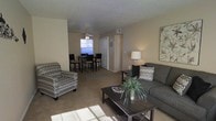 This single-story residence offers an open-concept layout that seamlessly connects the living and dining areas, creating a cohesive and functional living environment. The living area is comfortably sized, featuring wall-to-wall carpeting in a warm neutral tone that extends throughout the space. A dark wood coffee table anchors the seating area, accompanied by a charcoal-toned sofa and a patterned accent chair that add visual interest without overwhelming the understated palette. Decorative throw pillows and a potted plant contribute warmth to the arrangement.

The dining area is positioned toward the rear of the open space and benefits from a window that draws in natural light, brightening the room. A ceiling fan with integrated lighting serves both the dining and transitional zones. Dark wood dining chairs surround a matching table, maintaining a consistent design aesthetic.

The walls are finished in a soft beige tone, complemented by a textured ceiling. A closet with paneled doors is visible along the back wall, offering practical storage. The overall ambiance is calm and neutral, providing a versatile backdrop well-suited to a variety of furnishing styles.
