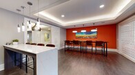 This thoughtfully designed single-floor amenity space combines vibrant aesthetics with functional gathering areas, creating an inviting environment for socializing and collaboration. The entrance lobby sets a welcoming tone with floor-to-ceiling glass doors and windows that flood the space with natural light, framing outdoor greenery views. A bold, patterned accent wall complements a seating arrangement of warm-toned sofas, teal accent chairs, and a colorful area rug, all beneath an elegantly coffered, backlit ceiling. The fully equipped kitchen is a standout feature, showcasing a striking turquoise accent wall, dark wood cabinetry, stainless steel appliances, and a generous quartz-topped island with bar seating — ideal for casual gatherings. A dedicated bar and display cabinet area with glass-front shelving and quartz countertops provides additional serving space. The dining and collaborative zone features high-top tables with leather bar stools set against a bold orange accent wall, anchored by large pendant lights and a vibrant cityscape artwork. Curated paintings line the connecting hallways, and a decorative lattice partition elegantly separates zones while maintaining an open flow. Durable hardwood-style flooring runs throughout, tying together the space's modern, energetic character.