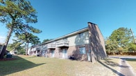 Located at 12012 Middleground Rd in Savannah, Georgia, this single-floor residence is part of a well-established brick building that blends classic construction with practical design. The exterior showcases a traditional brick façade with a neutral upper paneling, lending the structure a timeless, sturdy appearance. Each unit benefits from its own private entrance at ground level, providing convenience and a sense of individual access. Upper-level units feature open balconies with wood railings, offering outdoor space that overlooks a generous, open lawn. The surrounding grounds are dotted with mature pine trees, creating a natural, shaded environment that enhances the overall sense of calm and privacy. The property is set back from the street, allowing for ample open space and greenery that frames the building on multiple sides. Practical amenities such as exterior HVAC units are neatly positioned along the building's perimeter. The architecture, while straightforward, is solidly built and well-maintained, reflecting the established character of the Savannah, GA area. The single-floor layout provides ease of access, making it a practical and comfortable living option in a serene, tree-lined setting on Savannah's southside.