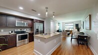 This thoughtfully designed 2-bedroom, 2-bathroom residence in Austin, TX offers a single-level layout with a seamless open-concept flow between the kitchen and living areas. The kitchen is a standout, featuring rich dark-wood cabinetry, stainless steel appliances, a tiled backsplash, and a central granite-topped island with a built-in sink and pendant lighting overhead. Warm hardwood flooring extends throughout the main living areas, complementing the soft, neutral color palette. The living room offers a comfortable, airy atmosphere with natural light streaming in through large windows. Both bedrooms are carpeted and well-proportioned, with the primary bedroom offering ample space and outdoor views through large windows fitted with roller shades. The second bedroom also benefits from multiple windows and a ceiling fan. The bathrooms feature clean, modern tile work, including a soaking tub with a tiled surround in warm taupe tones. A hallway connects the private and shared spaces efficiently. Recessed lighting and pendant fixtures throughout add to the polished, contemporary feel of this Austin residence.