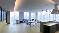 This contemporary single-floor space offers a refined and versatile environment designed for both collaboration and relaxation. The most striking feature is the expansive floor-to-ceiling glass curtain wall that wraps around the perimeter, flooding the interior with abundant natural light and framing impressive panoramic views of the surrounding urban skyline.

The open-plan layout is thoughtfully divided into distinct functional zones. A casual lounge area is anchored by a plush navy blue sofa, complementary armchairs, and a low coffee table set atop a decorative area rug, creating an inviting space for informal gatherings. Adjacent to this is a dining and meeting area furnished with sleek dark rectangular tables and modern white molded chairs, ideal for group work sessions or shared meals.

A kitchenette counter runs along one side, providing a practical service area that supports the social functions of the space. The light hardwood flooring extends throughout, lending warmth and continuity to the overall design. Overhead, a smooth cove ceiling with recessed lighting and a single pendant fixture adds an elegant architectural touch, contributing to the space's polished, professional ambiance. The overall atmosphere is bright, airy, and modern — well-suited for a high-rise amenity lounge or collaborative work environment.