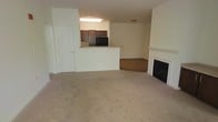 This single-floor space features an open-concept layout that connects the living area and kitchen seamlessly. The living room is finished with wall-to-wall neutral carpet, creating a warm and comfortable atmosphere. A white-mantled fireplace with a black insert serves as a focal point along one wall, adding both character and functional warmth to the space.

The kitchen is positioned at a slightly elevated counter level, separated from the living area by a half-wall pass-through that doubles as a breakfast bar. Dark espresso-toned cabinetry lines the kitchen walls, contrasting with the lighter walls and countertops. A black refrigerator is visible within the kitchen, and overhead fluorescent lighting illuminates the space efficiently.

Adjacent to the kitchen pass-through, a separate area with hardwood-style flooring can be seen, likely serving as a dining space. The overall color palette is neutral, with cream-toned walls throughout. A white paneled door near the kitchen provides access to a utility or storage area. The layout is practical and straightforward, maximizing usable space while maintaining an open, airy feel throughout the main living areas.