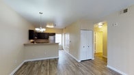 This single-story space features an open-concept design that seamlessly connects the kitchen and living areas, creating a comfortable and functional layout. The flooring throughout is finished in warm wood-style planks, lending a cohesive and inviting feel to the interior.

The kitchen is outfitted with dark espresso-toned cabinetry that contrasts elegantly against light granite-style countertops. A peninsula counter provides additional workspace and serves as a natural divider between the kitchen and the main living area. A stainless steel refrigerator is visible, and natural light filters in through a window above the sink area. An elegant chandelier-style pendant light hangs above the peninsula, adding a decorative touch.

The open living area is generously proportioned, with neutral warm-toned walls and crisp white trim that brighten the space. A set of white bifold closet doors offers convenient storage. A hallway leads toward additional rooms, including what appears to be a bedroom and bathroom, each accessible through paneled doors. The layout flows naturally from one area to the next, making the space feel organized and well-designed throughout.