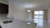 This single-floor space showcases an open-concept layout that seamlessly connects the kitchen and living area, creating a bright and functional environment. The kitchen is equipped with polished granite countertops featuring subtle flecks of color, a stainless steel sink with a modern arc faucet, and a peninsula-style counter that serves as a natural divider between the cooking and living spaces. A stylish pendant light with a brushed nickel finish hangs above, providing focused illumination and a refined decorative touch. The floors throughout are finished in large-format light-toned tiles, contributing to a clean and cohesive aesthetic. The living area flows naturally toward a full-width sliding glass door that opens onto a private balcony enclosed by a metal railing, offering pleasant outdoor views of greenery and what appears to be a water feature in the distance. The textured ceiling adds subtle visual depth, while the neutral wall tones provide a versatile backdrop suitable for a variety of interior styles. Natural light filters generously through the balcony access point, enhancing the overall sense of openness throughout the space.