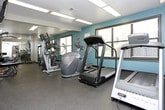 Located at 6097 Memorial Dr in Stone Mountain, Georgia, this fitness center offers a practical and well-equipped space for residents or members seeking a convenient workout facility. The single-floor layout is thoughtfully organized to accommodate a variety of exercise needs, from cardio to strength training.

The main workout area features durable rubber flooring throughout, providing both safety and comfort during exercise. Overhead fluorescent panel lighting ensures the space is brightly and evenly illuminated. The room is finished with a clean combination of white and teal accent walls, lending the space a modern, energetic feel.

Cardio equipment includes Matrix treadmills and elliptical machines, positioned to take advantage of natural light from multiple windows fitted with horizontal blinds. A wall-mounted flat-screen television adds entertainment options for those logging longer cardio sessions.

The strength training section is well-stocked with a multi-function cable/pull-up tower, adjustable benches, dumbbells, and additional resistance machines, offering a comprehensive range of exercise options. Full-length mirrors along one wall enhance the sense of openness and allow users to monitor their form.

Multiple entry and exit points, including a windowed door leading to the exterior, contribute to the room's accessibility and ventilation. A fire extinguisher is visibly mounted for safety compliance. Overall, this fitness center provides a functional and inviting environment for a full-body workout.