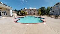 Located at 111 Coopers Ridge Blvd in Ladson, South Carolina, this outdoor amenity area serves as a central communal gathering space within the residential community. The centerpiece is a generously sized freeform swimming pool with a classic brick-bordered edge and clear blue water, marked for shallow entry at 3 feet. The pool is complemented by an expansive concrete sundeck that surrounds the water on all sides, providing ample room for lounging and recreation. Numerous lounge chairs are arranged along the perimeter, offering plentiful seating for residents to enjoy the South Carolina sunshine. A covered pavilion with white columns and a brick base provides a shaded retreat at the edge of the deck, ideal for relaxing out of direct sun. Access to the pool area is controlled via a secured entry door fitted with a keypad and utility connections mounted to the building's exterior lap siding. The surrounding buildings are clad in neutral-toned vinyl lap siding with dark green architectural accents and feature multi-story balconies that overlook the pool courtyard. Mature trees frame the backdrop of the community, adding a natural, landscaped setting to the outdoor environment. The entire space is designed to encourage outdoor leisure and community engagement in a well-maintained residential setting.