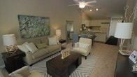This single-floor residence offers an open-concept design that seamlessly connects the living, dining, and kitchen areas into one cohesive and functional space. The living area is anchored by plush, neutral-toned seating arranged around a dark wood coffee table and accented by a chevron-patterned area rug, creating a comfortable and stylish focal point. A striking vaulted ceiling fitted with a ceiling fan and track lighting adds a sense of height and airiness throughout the main living zone. Adjacent to the living area, a dedicated dining space accommodates a table and upholstered seating, ideal for both casual meals and entertaining. The kitchen is positioned directly off the dining area and features light-colored cabinetry, countertop appliances, and a double sink, offering practical functionality within easy reach. Warm, neutral wall tones and soft carpeting throughout contribute to an inviting and cohesive ambiance. A large decorative mirror near the entryway enhances the sense of space, while a floral canvas print adds a touch of color and character to the living area. The overall design balances comfort and practicality in a bright, well-organized layout.